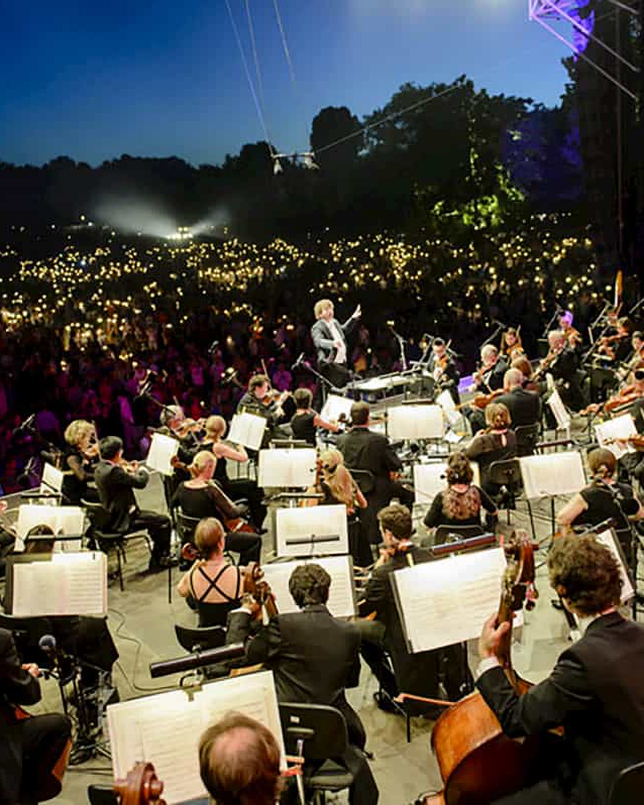 An orchestra playing open air at dusk in front of a sea of lights